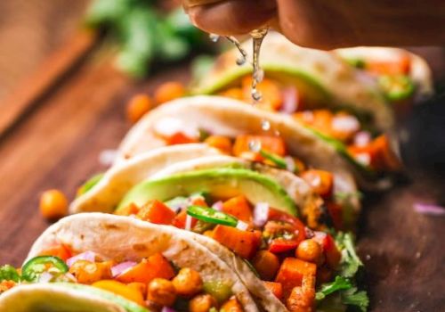 The image shows multiple tacos filled with chickpeas, vegetables, and avocado slices, with a hand squeezing lime juice over them.