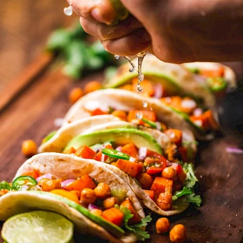 The image shows multiple tacos filled with chickpeas, vegetables, and avocado slices, with a hand squeezing lime juice over them.
