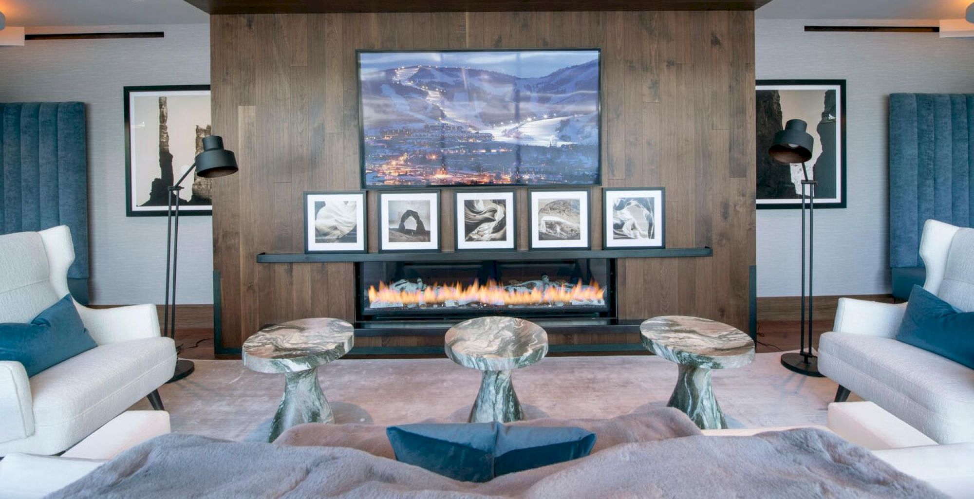 A modern living room with a fireplace, artwork on the mantle, a large TV above, white chairs, and green stone tables in front.