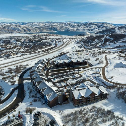 An aerial view of a snow-covered resort area with buildings, winding roads, and surrounding mountains under a blue sky.