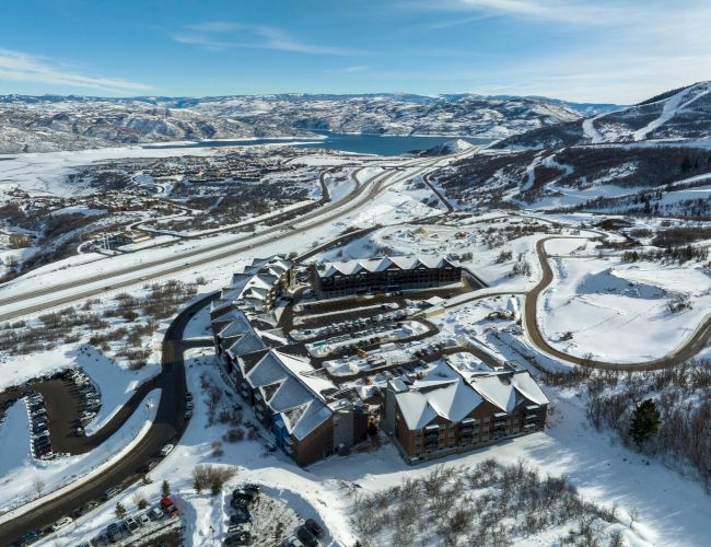 An aerial view of a snow-covered resort area with buildings, winding roads, and surrounding mountains under a blue sky.