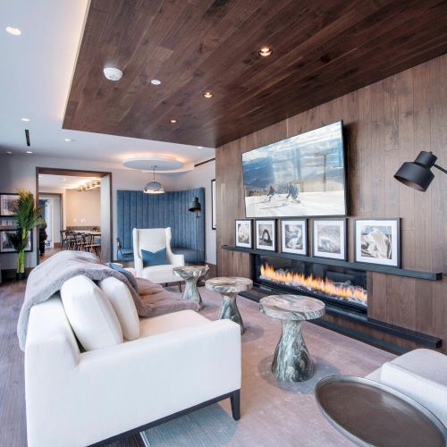 A modern living room with large windows, white sofas, a TV, a fireplace, and artistic decor on the wooden wall.