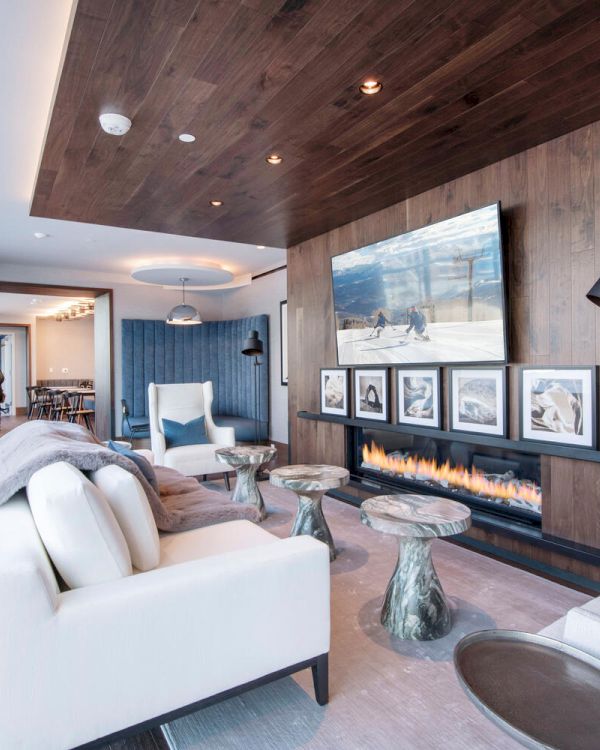 A modern living room with large windows, white sofas, a TV, a fireplace, and artistic decor on the wooden wall.