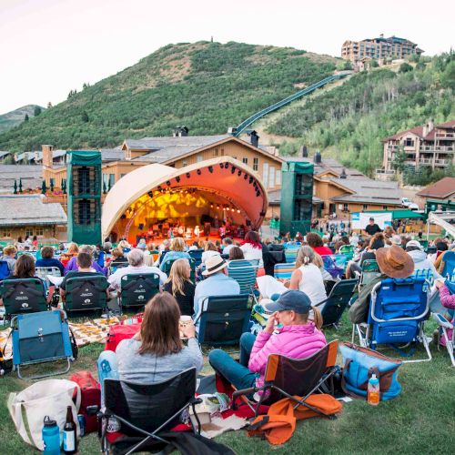 People gather on lawn chairs watching an outdoor stage concert in a hillside park, with a bright semicircular green stage and mountains in the background.