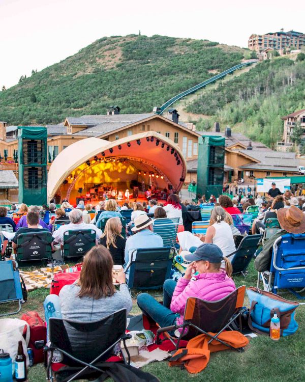 People gather on lawn chairs watching an outdoor stage concert in a hillside park, with a bright semicircular green stage and mountains in the background.