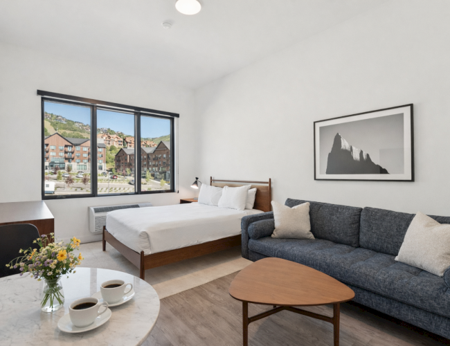 A bright hotel room with a large window, a bed, a gray sofa, a wooden coffee table, and a small round table with coffee and flowers, ending this sentence.