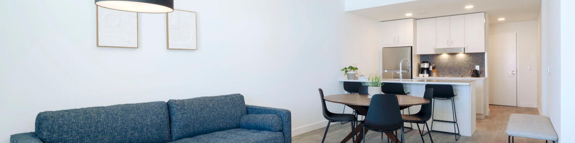 A modern living room with a blue sofa, a wooden coffee table, and a kitchen in the background featuring white cabinets and a dining area.