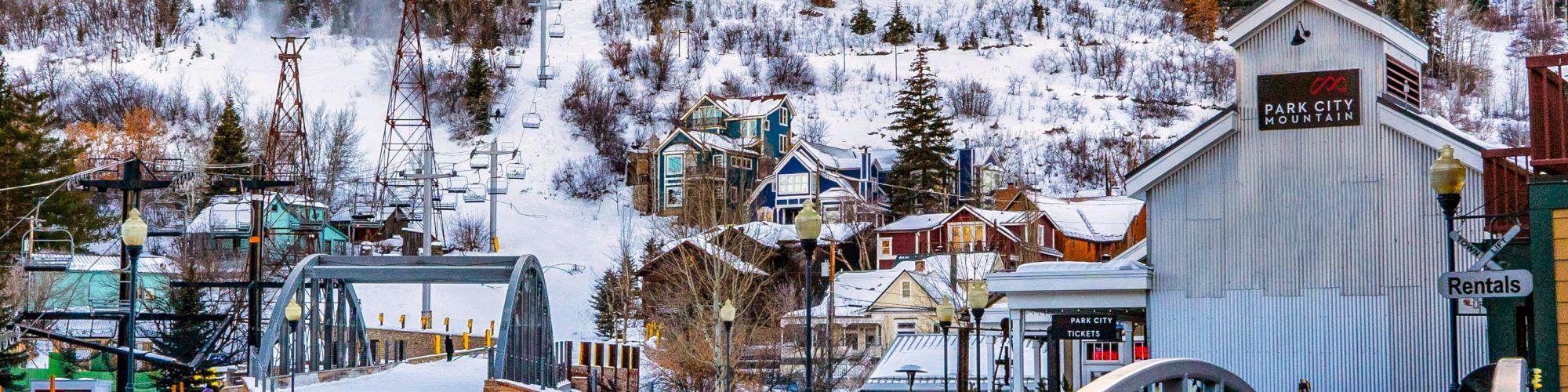 A snowy mountain with ski trails runs behind a picturesque town with colorful buildings, a bridge in the foreground, and a vibrant winter atmosphere.