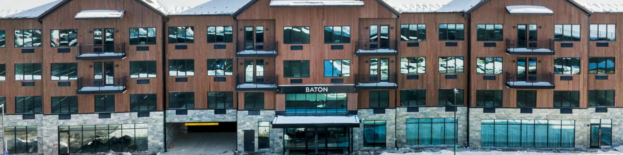 A large, multi-story building with a wooden facade, featuring multiple windows and a parking lot in the front, partially covered in snow.