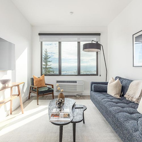 A modern living room with a blue sofa, wall-mounted TV, large window, floor lamp, wooden console table, and minimalist decor, overlooking a scenic view.