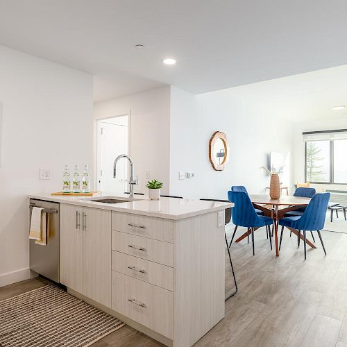 A modern open-concept kitchen and living area with a dining table, blue chairs, a sofa, large window, minimalist decor, and light wood flooring.