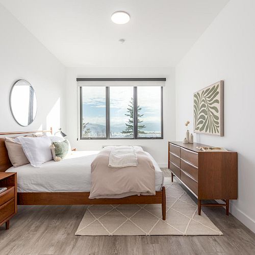 A minimalist bedroom with a bed, nightstand, lamp, dresser, abstract artwork, and a window showing an outdoor view.