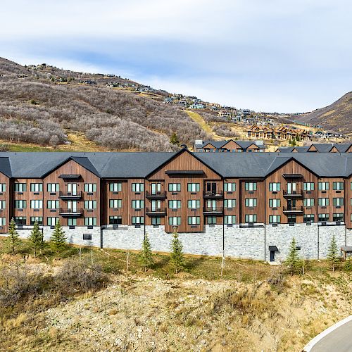 The image shows a large multi-story building with a wood and stone facade situated in a hilly area with sparse trees and additional buildings in the background.