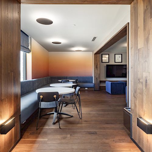 A modern room with wooden floors, tables, chairs, a sofa, and gradient wall art. The room is well-lit with ceiling lights and natural light from a window.