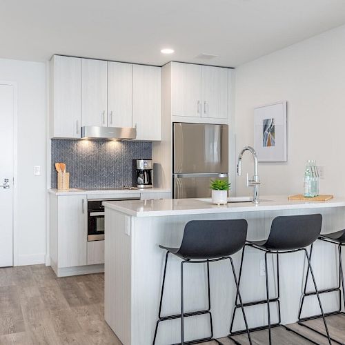 A modern kitchen with white cabinets, stainless steel appliances, an island with a sink, and three black bar stools.