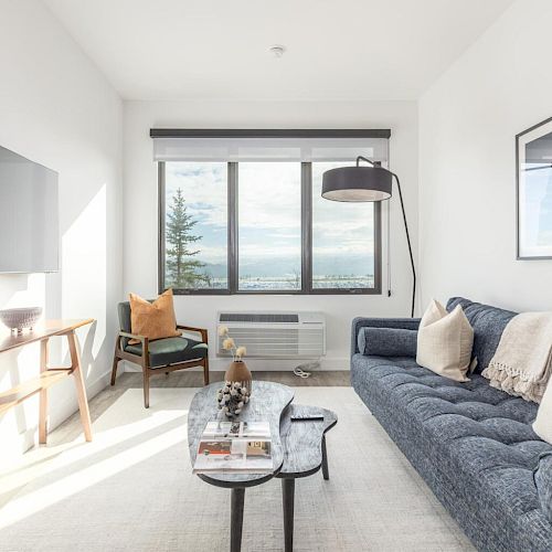 A modern living room with a couch, a coffee table, a wall-mounted TV, a large window with a view, and a floor lamp is shown.