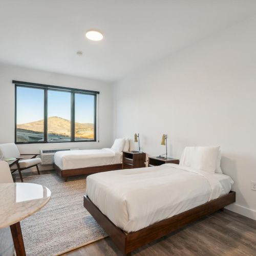 The image shows a bedroom with two single beds, a large window, a TV, and a round table, decorated in a minimalist style.