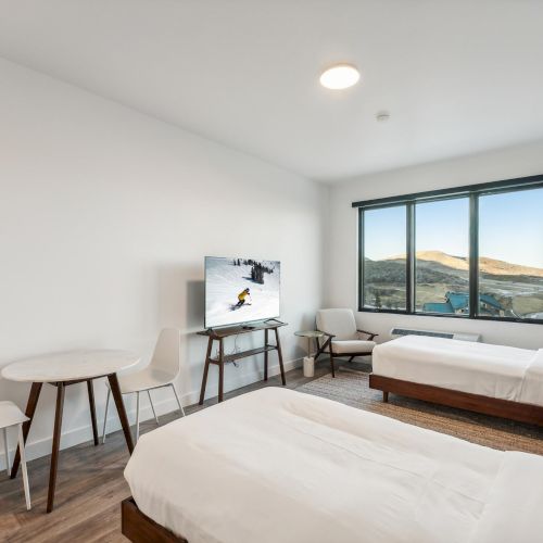 The image shows a modern room with two beds, a table with chairs, a TV, and a window offering a scenic view of hills and sky outside.