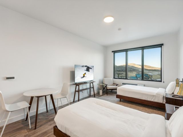 A modern hotel room with two beds, a TV, a table, and chairs, and a large window with a view.