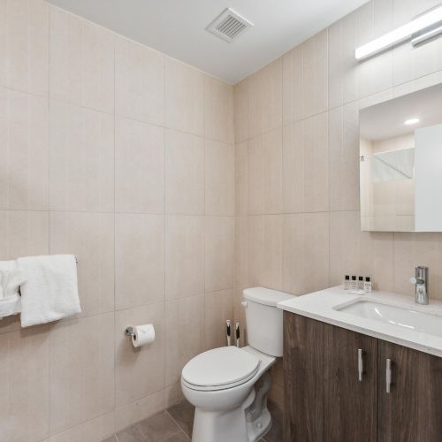 A clean bathroom with a toilet, vanity sink, mirror, and towel rack on a tiled wall. Bright lighting and minimal decor complete the space.