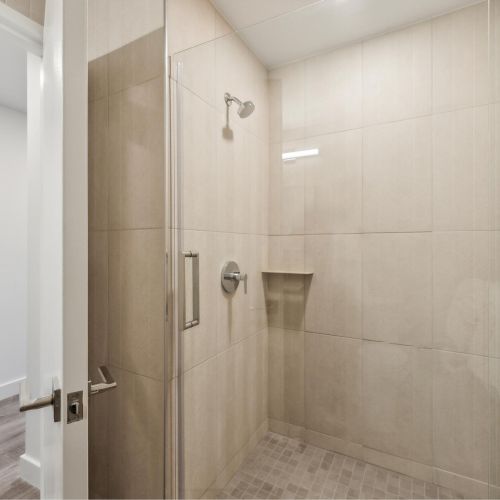 The image shows a modern tiled shower with glass doors, featuring a wall-mounted showerhead and a small recessed shelf for toiletries.