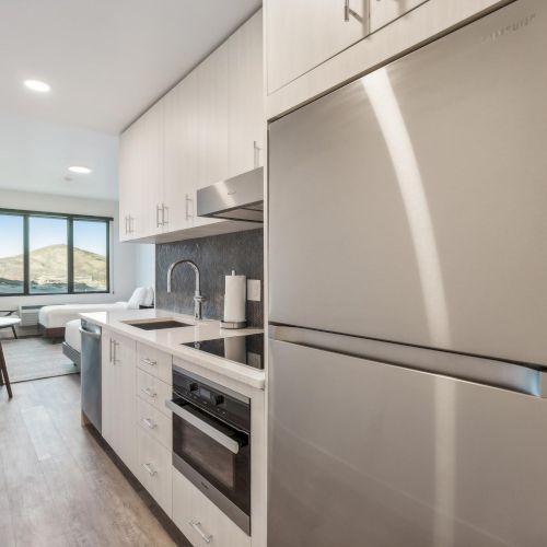 A modern, narrow kitchen with white cabinets, stainless steel appliances, and wood flooring leads to a living area with a large window.