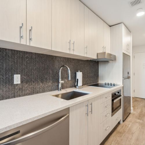 A modern kitchen with light wood cabinets, dark herringbone backsplash, stainless steel appliances, and a quartz countertop is shown.