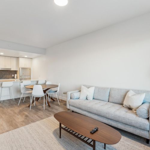 A modern living room with gray sofa, coffee table, floor lamp, and a kitchen area with a dining table and chairs.