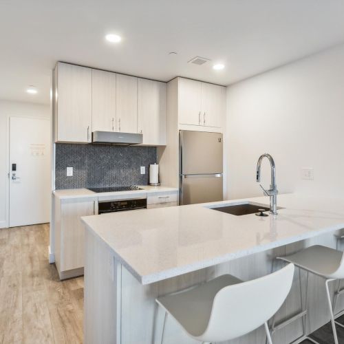 A modern kitchen with white cabinets, a stainless steel fridge, an island with barstools, and wood flooring, under bright lighting.