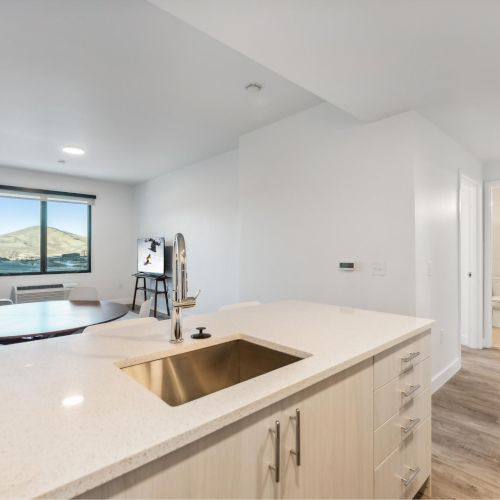 A modern open-concept apartment with a kitchen, living area, and a window view. There's a hallway leading to a bathroom and another room.