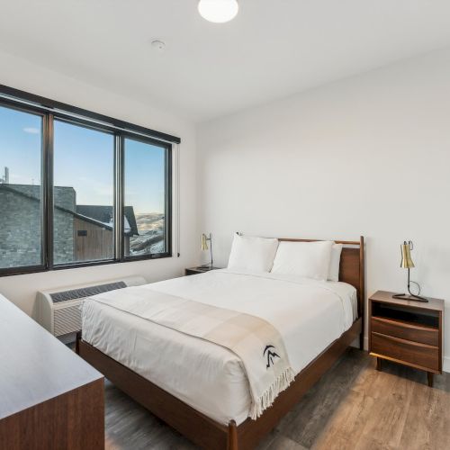 A modern bedroom with a bed, white bedding, two side tables with lamps, a dresser, and a large window showing an outside view.