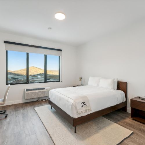 A minimalist bedroom with a bed, side table, desk, and chair. Large window with a view of hills. The floor is wooden, and the walls are white.