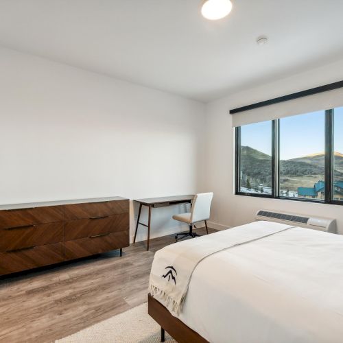 A minimalist bedroom with a bed, wooden dresser, desk, chair, and large window showcasing a scenic view.