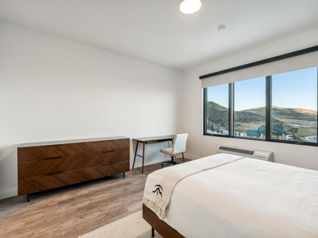 A hotel room with a large window, a bed, a desk with a chair, and a dresser.