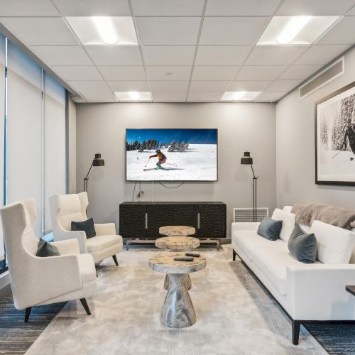 A modern living room with chairs, a sofa, a wall-mounted TV, and a skiing photo. Decor is minimalistic with neutral colors.