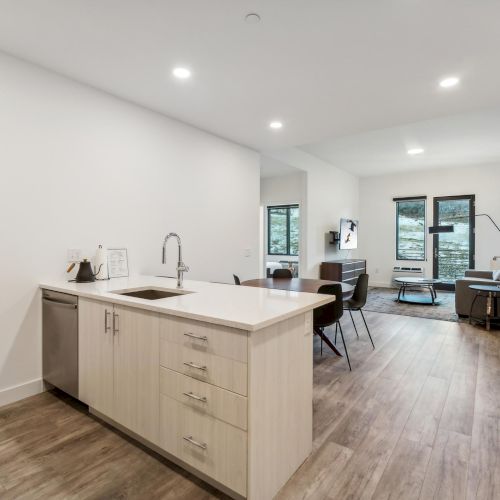 A modern open-plan kitchen and living area with wooden floors, a central island, dining table, artwork, and large windows.