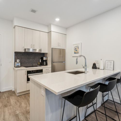 This image shows a modern kitchen with light cabinets, a fridge, a sink on a white island, black chairs, and wood flooring.