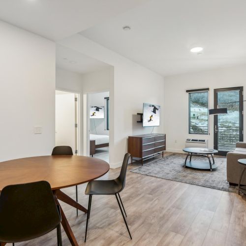 A modern living room with wood flooring, a round table with chairs, sofa, TV, and large windows letting in natural light.