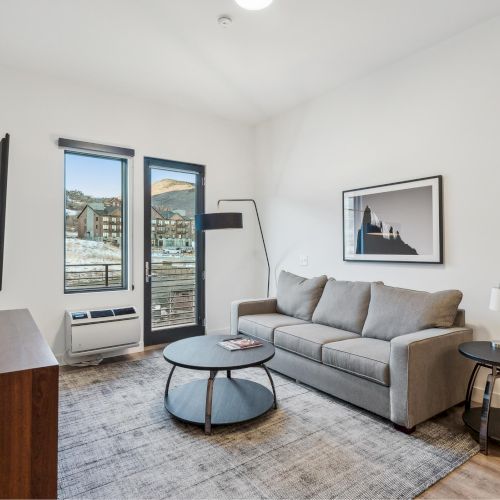 A modern living room with a gray couch, round tables, wall-mounted TV, floor lamp, and artwork. Large windows show a scenic outdoor view.