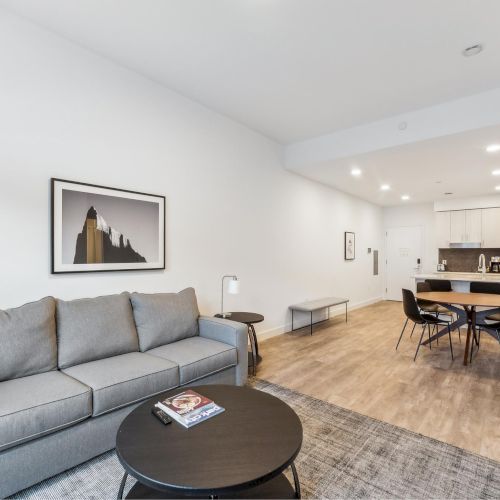 A modern living room with a gray sofa, round table, artwork, and open kitchen with dining table and chairs on wooden floors.