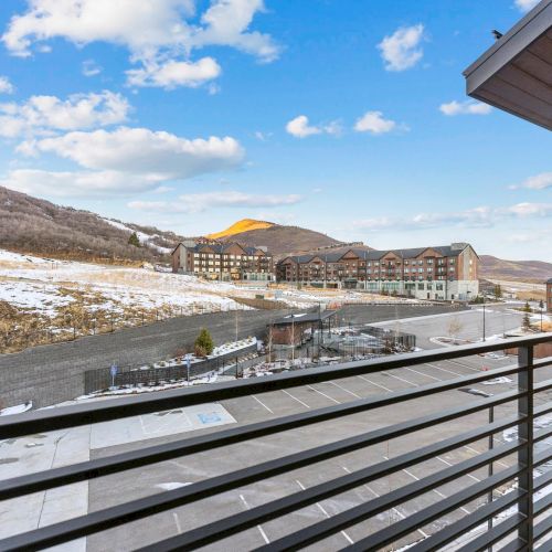 A scenic view from a balcony with a mountain landscape, buildings, and a partly snow-covered ground under a blue sky with clouds.