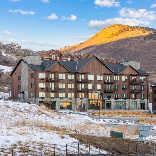 A large wooden lodge-style building is set against a mountainous, snowy landscape under a blue sky with clouds.