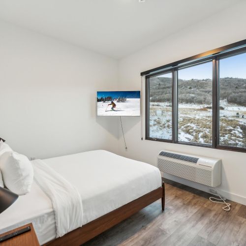 A bedroom with a bed, wall-mounted TV, large window showing a snowy landscape, and a heater below the window.