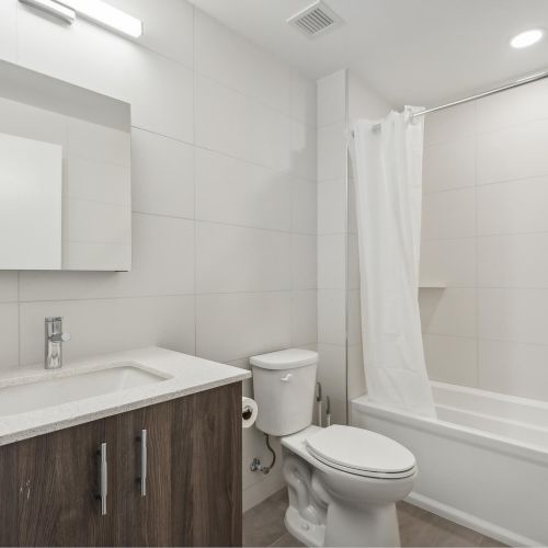 A clean, modern bathroom with a sink, mirror, toilet, and bathtub with a shower curtain in a neutral-toned setting.