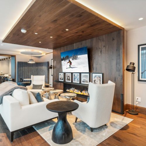 A modern living room with white chairs, a wooden ceiling, TV, artwork, and decor. A dining area is visible in the background.