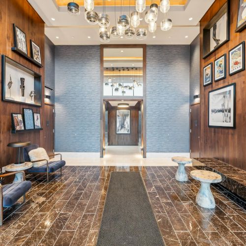 A modern lobby with art-adorned walls, plush seating, and stylish lighting fixtures, featuring a central elevator at the end of the corridor.