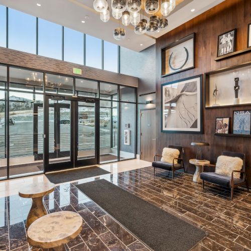 A modern lobby with decorative lighting, art on the walls, and sleek furniture includes chairs and tables on a polished floor, leading to glass doors.