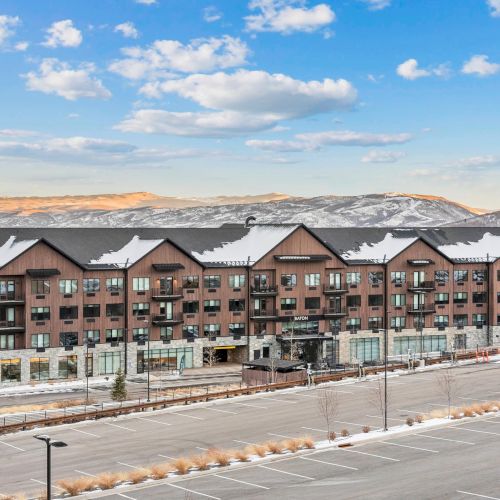 The image shows a large, multi-story building with a snowy roof, set against a mountainous landscape and a mostly empty parking lot in the foreground.