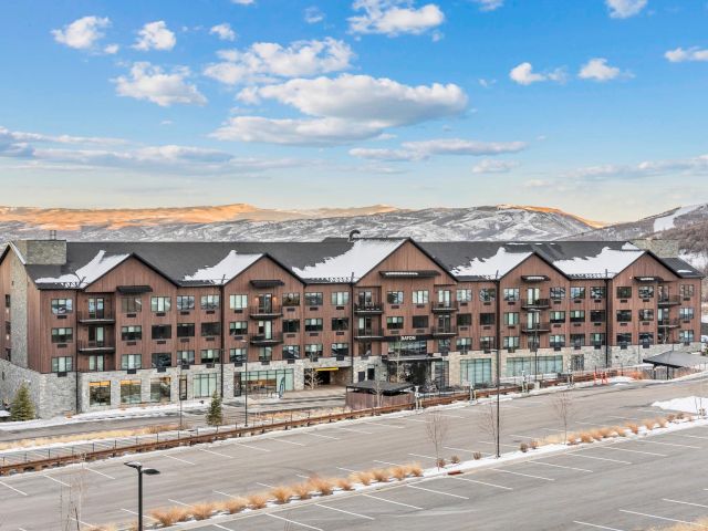 A multi-story brown and gray building with snow on its roof, set against a backdrop of mountains and a blue sky with clouds.
