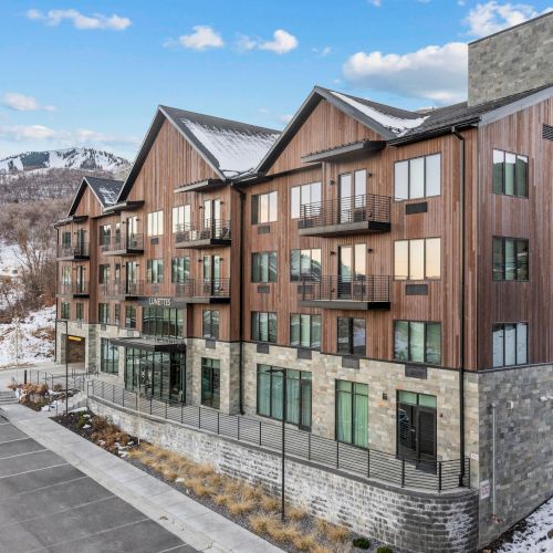 The image shows a modern, multi-story building with wood and stone exterior, surrounded by a snowy landscape and mountains in the background.